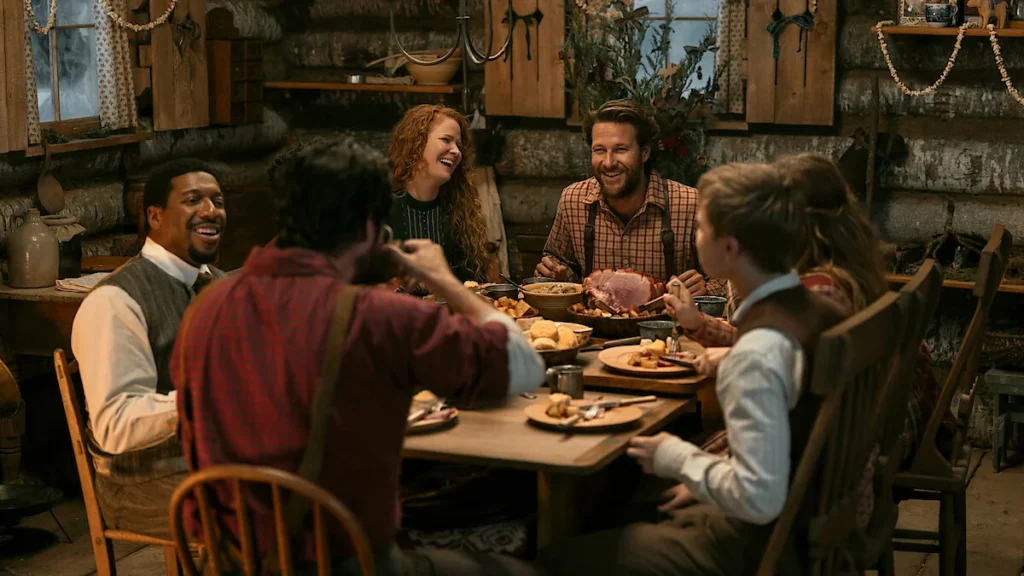 Personagens de Uma Casa na Pradaria reunidos ao redor de uma mesa durante uma refeição na cabana.